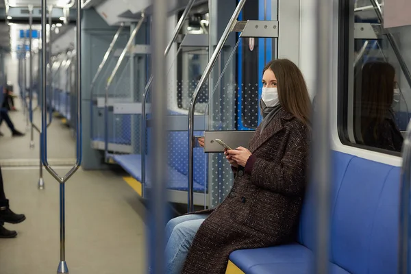 A woman in a medical face mask to avoid the spread of coronavirus is sitting holding a smartphone in a modern subway car. A girl in a surgical mask against COVID-19 is taking a ride on a train.