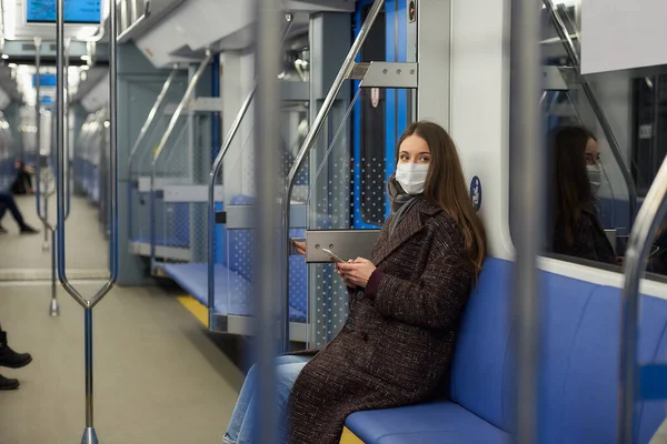 A woman in a medical face mask to avoid the spread of coronavirus is sitting holding a smartphone in a modern subway car. A girl in a surgical mask against COVID-19 is taking a ride on a metro train.