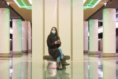 COVID 'nin yayılmasını önlemek için tıbbi maskeli bir kadın akıllı telefonuyla oturmuş metro trenini bekliyor. Cerrahi maske takan uzun saçlı bir kız metroda sosyal mesafeyi koruyor..