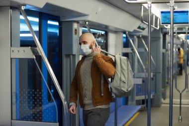 Koronavirüsün yayılmasını önlemek için maskeli sakallı bir adam metroya binerken gri bir sırt çantası takıyor. Cerrahi maskeli kel bir adam trende sosyal mesafeyi koruyor..
