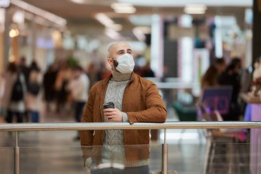 Coronavirus 'un yayılmasını önlemek için maske takan bir adam alışveriş merkezinde beklerken elinde bir fincan kahve tutuyor. Cerrahi maskeli kel bir adam sosyal mesafeyi koruyor..