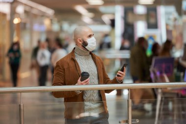 Yüz maskesi takmış sakallı bir adam bakınıyor ve alışveriş merkezinde elinde akıllı bir telefon ve bir fincan kahve tutuyor. Cerrahi maskeli kel bir adam sosyal mesafeyi koruyor..