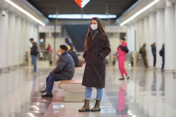 A full-length photo of a woman in a medical face mask to avoid the spread of coronavirus who is waiting for a train in the center of a metro station. Girl in a surgical mask is keeping social distance