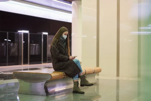 一名戴着医疗面罩以避免考罗纳威病毒传播的女性坐在地铁平台上 并使用智能手机 一个戴口罩的女孩在地铁里与外界保持着距离 — 图库照片
