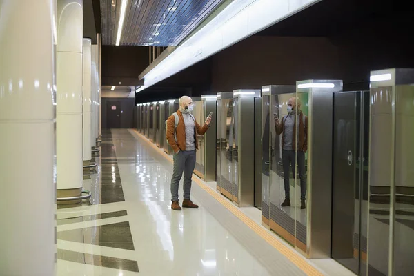 A man in a face mask to avoid the spread of coronavirus is using a smartphone while waiting for a train at the subway platform. A bald guy in a surgical mask is keeping social distance.