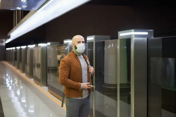 A man in a medical face mask is holding a smartphone and staring to the side while waiting for a train at the subway platform. A bald guy in a surgical mask is keeping social distance.