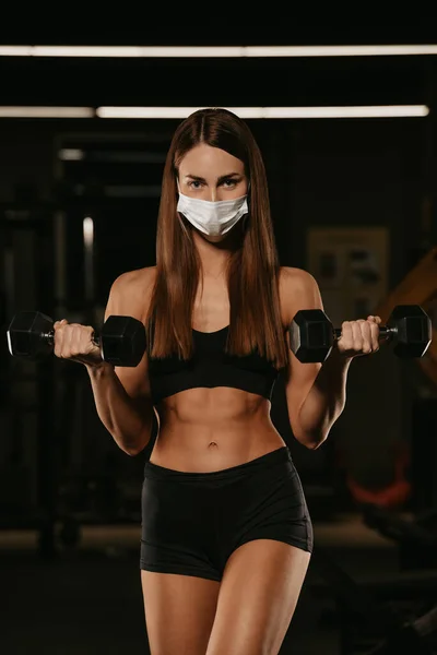 A fit woman in a face mask to avoid the spread of coronavirus is doing bicep curls with dumbbells. A sporty girl in a surgical mask is posing during the arms workout in a gym.