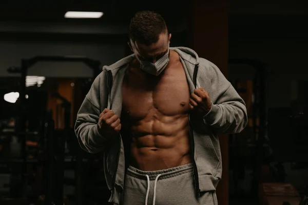 A bodybuilder in a face mask to avoid the spread of coronavirus is opening his zipped hoodie to demonstrate his athletic physique. A sporty guy in a surgical mask is posing after a workout in a gym.