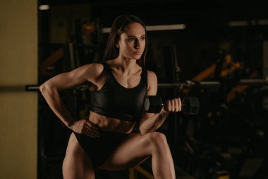 Sportif bir kadın spor salonunda dirseğiyle halter kıvırıyor. Kaslı esmer bir kız siyah bluz giyer ve kısa şort giyer..