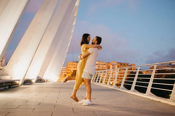 A man with a beard is hugging the waist of his girl in a yellow dress on a white bridge in Valencia. A couple of tourists in full length on a date in the warm evening.