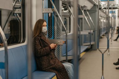 Coronavirus 'un yayılmasını önlemek için tıbbi maske takan bir kadın modern bir metro vagonunda elinde akıllı bir telefonla oturuyor. COVID-19 'a karşı cerrahi maskeli bir kız trene biniyor..