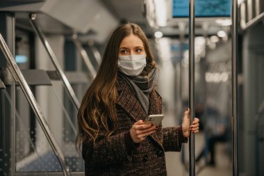 Coronavirus 'un yayılmasını önlemek için maske takan kadın modern bir metro vagonunda ayakta duruyor ve yan tarafa bakıyor. COVID-19 'a karşı cerrahi maske takan bir kız metro trenine biniyor.