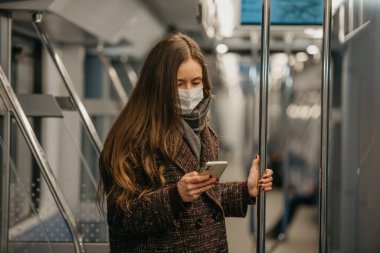 Coronavirus 'un yayılmasını önlemek için tıbbi maske takan bir kadın modern bir metro vagonunda ayakta duruyor ve akıllı telefon kullanıyor. Cerrahi maskeli bir kız trende cep telefonuyla haberlere çıkıyor..