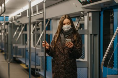Coronavirus 'un yayılmasını önlemek için tıbbi maske takan bir kadın boş bir metro vagonunda akıllı telefon kullanıyor. Cerrahi maskeli bir kız trende cep telefonuyla haberlere çıkıyor..