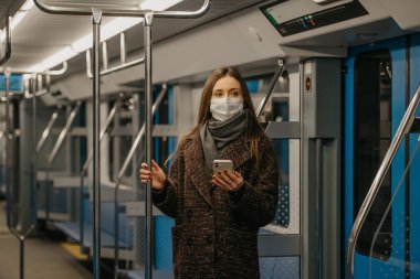 Coronavirus 'un yayılmasını önlemek için tıbbi maskeli bir kadın modern bir metro vagonunda ayakta duruyor ve elinde bir akıllı telefon tutuyor. COVID-19 'a karşı ameliyat maskeli bir kız metroya biniyor..
