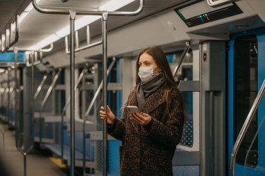 Coronavirus 'un yayılmasını önlemek için tıbbi maske takan bir kadın modern bir metro vagonunda duruyor ve elinde telefon tutuyor. COVID-19 'a karşı cerrahi maske takan bir kız metro trenine biniyor..