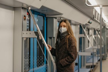 Coronavirus 'un yayılmasını önlemek için tıbbi maske takan bir kadın kapıların yanında duruyor ve modern bir metro vagonunun kenarına bakıyor. Cerrahi maskeli bir kız metro trenine biniyor..