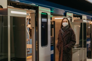 Koronavirüsün yayılmasını önlemek için tıbbi maske takan bir kadın modern metro vagonunun yanında bekliyor. Cerrahi maskeli bir kız metro istasyonunda sosyal mesafeyi koruyor..