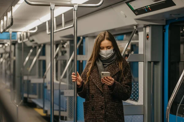 A woman in a medical face mask to avoid the spread of coronavirus is standing and using a smartphone in an empty subway car. A girl in a surgical mask is scrolling news on her cellphone on a train.