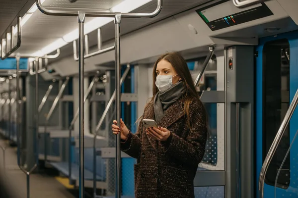 A woman in a medical face mask to avoid the spread of coronavirus is standing and holding a phone in a modern subway car. A girl in a surgical mask against COVID-19 is taking a ride on a metro train.