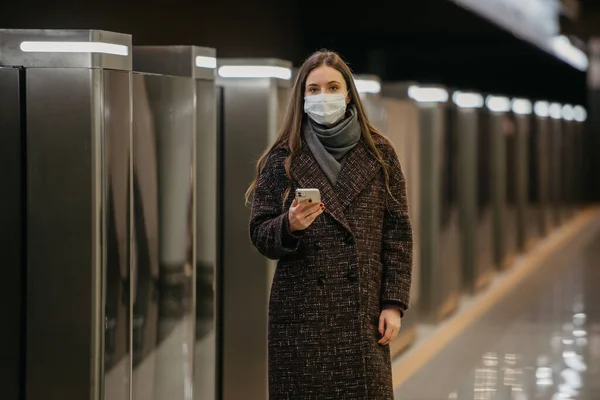 A woman in a medical face mask to avoid the spread of coronavirus is waiting for a train and holding a cellphone at the subway station. A girl in a surgical mask is keeping social distance in metro.