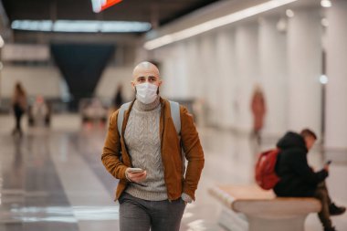 Tıbbi maskeli bir adam akıllı bir telefon tutuyor ve metro istasyonunun merkezinde tren beklerken yürüyor. Cerrahi maskeli kel bir adam sosyal mesafeyi koruyor..