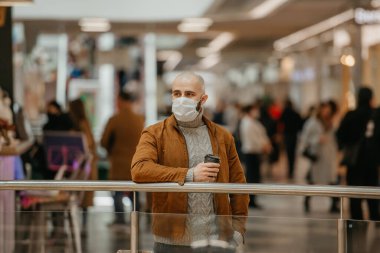 Coronavirus 'un yayılmasını önlemek için maske takan bir adam alışveriş merkezinde beklerken elinde bir fincan kahve tutuyor. Cerrahi maskeli kel bir adam sosyal mesafeyi koruyor..