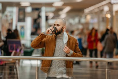 Alışveriş merkezinde bir adam tıbbi maskesini çıkarırken kahve içiyor. Kel bir adam sosyal mesafeyi koruyor..