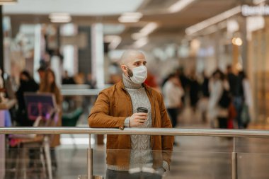 Coronavirus 'un yayılmasını önlemek için tıbbi maske takan bir adam alışveriş merkezinde beklerken elinde bir fincan kahve tutuyor. Cerrahi maskeli kel bir adam sosyal mesafeyi koruyor..