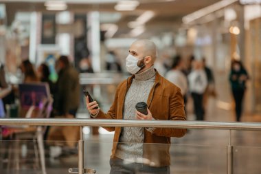 Yüz maskesi takmış sakallı bir adam bakınıyor ve alışveriş merkezinde elinde akıllı bir telefon ve bir fincan kahve tutuyor. Cerrahi maskeli kel bir adam sosyal mesafeyi koruyor..