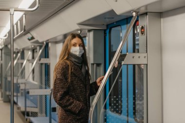 Coronavirus 'un yayılmasını önlemek için tıbbi maske takan bir kadın kapıların yanında duruyor ve modern bir metro vagonunun kenarına bakıyor. Cerrahi maskeli bir kız metro trenine biniyor..