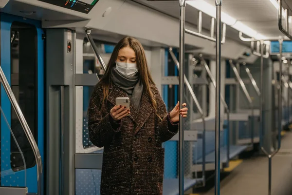 A woman in a medical face mask to avoid the spread of coronavirus is standing and using a smartphone in an empty subway car. A girl in a surgical mask is scrolling news on her cell phone on a train.