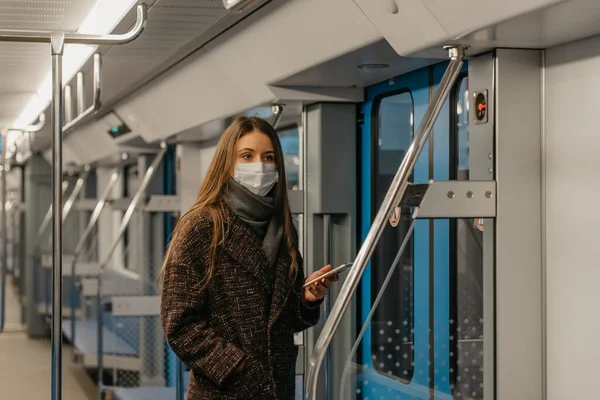 A woman in a medical face mask to avoid the spread of coronavirus is standing near doors and staring to the side in an empty subway car. A girl in a surgical mask is taking a ride on a metro train.