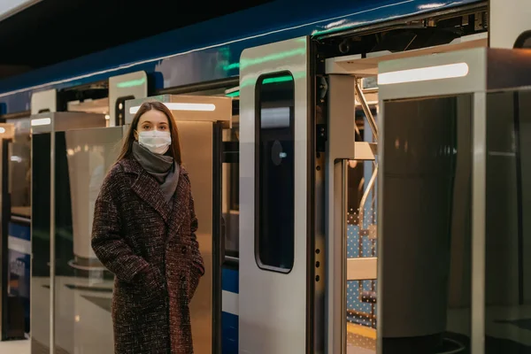 A woman in a medical face mask to avoid the spread of coronavirus is waiting near the modern subway car. A girl in a surgical mask is keeping social distance on a metro station.