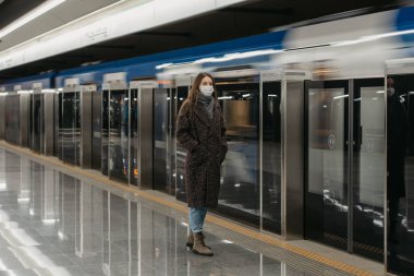 Metroda modern bir trenin gelmesini bekleyen Coronavirus 'un yayılmasını önlemek için tıbbi maske takan bir kadının tam boy fotoğrafı. Cerrahi maskeli kız sosyal mesafeyi koruyor..