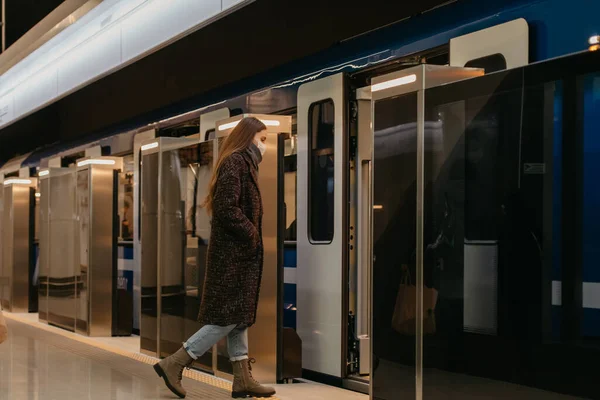 A full-length photo of a lady in a medical face mask to avoid the spread of coronavirus who is entering the modern subway car. A girl in a surgical mask is keeping social distance on a metro station.