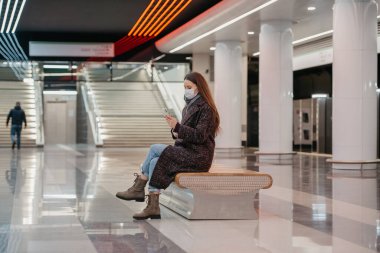 Tıbbi maske takan bir kadın metro istasyonunun ortasında oturuyor ve akıllı telefon kullanıyor. Cerrahi maske takan uzun saçlı bir kız metroda sosyal mesafeyi koruyor..