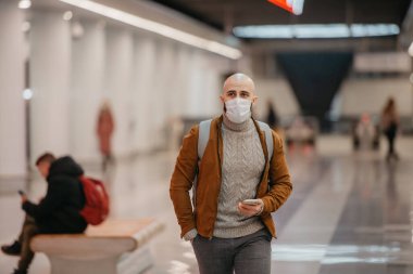 Tıbbi maskeli bir adam akıllı bir telefon tutuyor ve metro istasyonunun merkezinde tren beklerken yürüyor. Cerrahi maskeli kel bir adam sosyal mesafeyi koruyor..