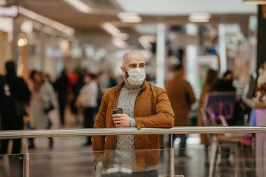 Coronavirus 'un yayılmasını önlemek için maske takan bir adam alışveriş merkezinde beklerken elinde bir fincan kahve tutuyor. Cerrahi maskeli kel bir adam sosyal mesafeyi koruyor..
