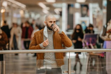 Bir adam alışveriş merkezinde kahve içerken tıbbi maske takıyor. Kel bir adam sosyal mesafeyi koruyor..
