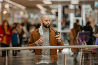 Bir adam alışveriş merkezinde kahve içerken poz veriyor ve tıbbi maske takıyor. Kel bir adam sosyal mesafeyi koruyor..