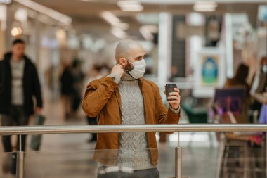 Sakallı bir adam alışveriş merkezinde kahverengi bir fincan kahve tutarken tıbbi yüz maskesi takıyor. Kel bir adam sosyal mesafeyi koruyor..