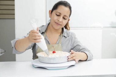 Hispanic woman in her patisserie decorating a cake - woman putting whipped cream on freshly baked cake - enterprising woman clipart