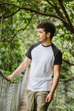 Ormanın ortasında asma köprüde yürüyen genç İspanyol bir adam - genç bir gezgin doğayı izlerken ve yolculuğun tadını çıkarırken - maceraperest bir adam