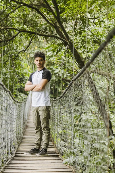 Young hispanic man walking on a suspension bridge in the middle of the forest - young traveler walking while observing nature and enjoying the ride - adventurous man