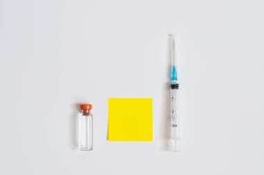Medical Injection, Plastic Syringe, Blank Yellow Piece Of Paper For Text And Bottle With Liquid On White Background With Copyspace. Top View.