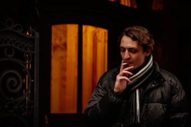 Yakışıklı, şık, genç hippi kışın sokakta sigara içip gözlerini kaçırıyor. Sıradan giyinmiş güzel bir adamın portresi. Klasik Avrupa şehri, arka planda ışıklar. Yumuşak odak.