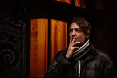 Yakışıklı, şık, genç hippi kışın sokakta sigara içip gözlerini kaçırıyor. Sıradan giyinmiş güzel bir adamın portresi. Klasik Avrupa şehri, arka planda ışıklar. Yumuşak odak.