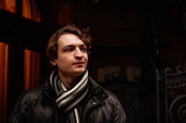 Sokakta kışlık kıyafetler giyen yakışıklı, şık, genç bir hippi. Ceket ve atkıyla modern saç stili olan güzel bir adamın portresi. Klasik Avrupa şehri, arka planda ışıklar. Yumuşak odak.