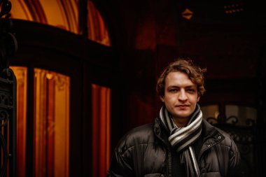 Sokakta kışlık kıyafetler giyen yakışıklı, şık, genç bir hippi. Ceket ve atkıyla modern saç stili olan güzel bir adamın portresi. Klasik Avrupa şehri, arka planda ışıklar. Yumuşak odak.